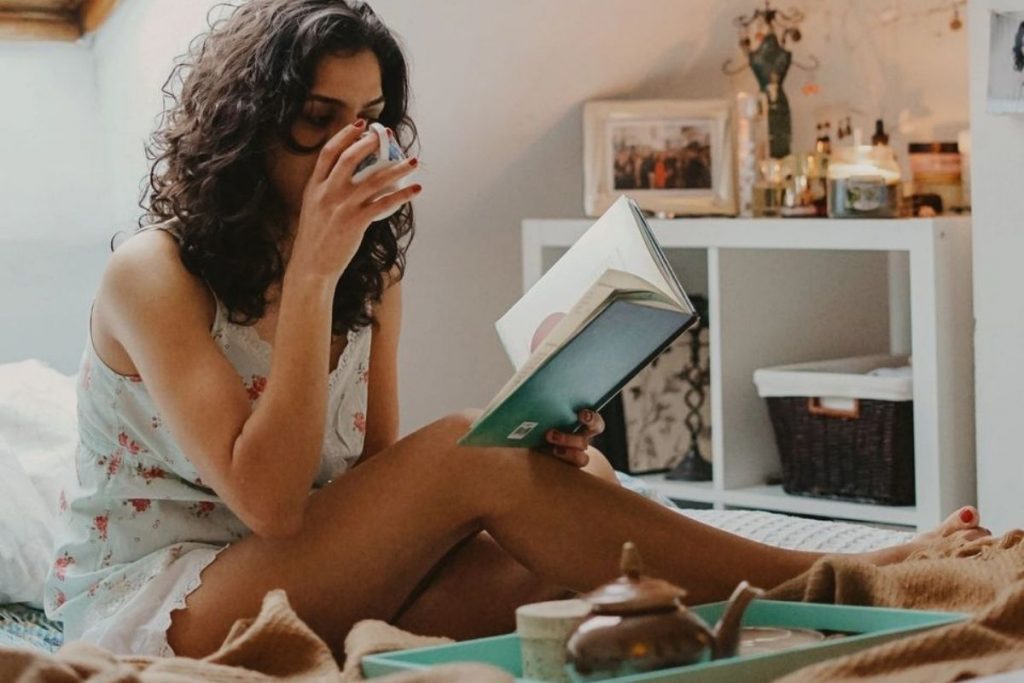 woman sitting on a bed with a cup of tea and a book taking care of herself