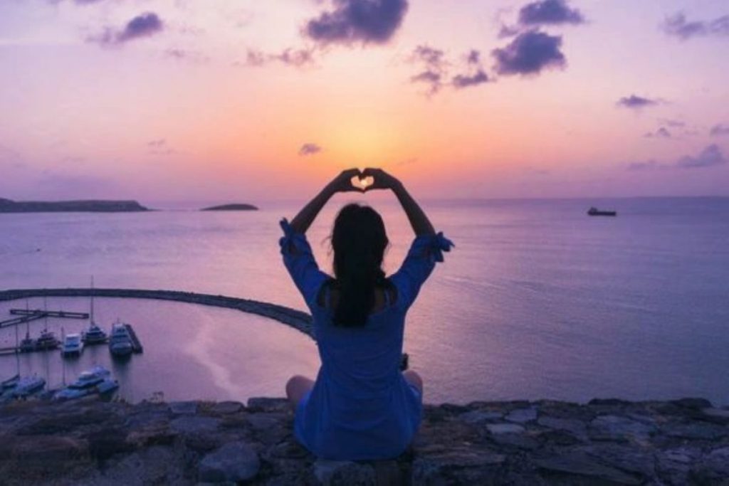 woman sitting on the shore doing yoga looking over a purple sunset