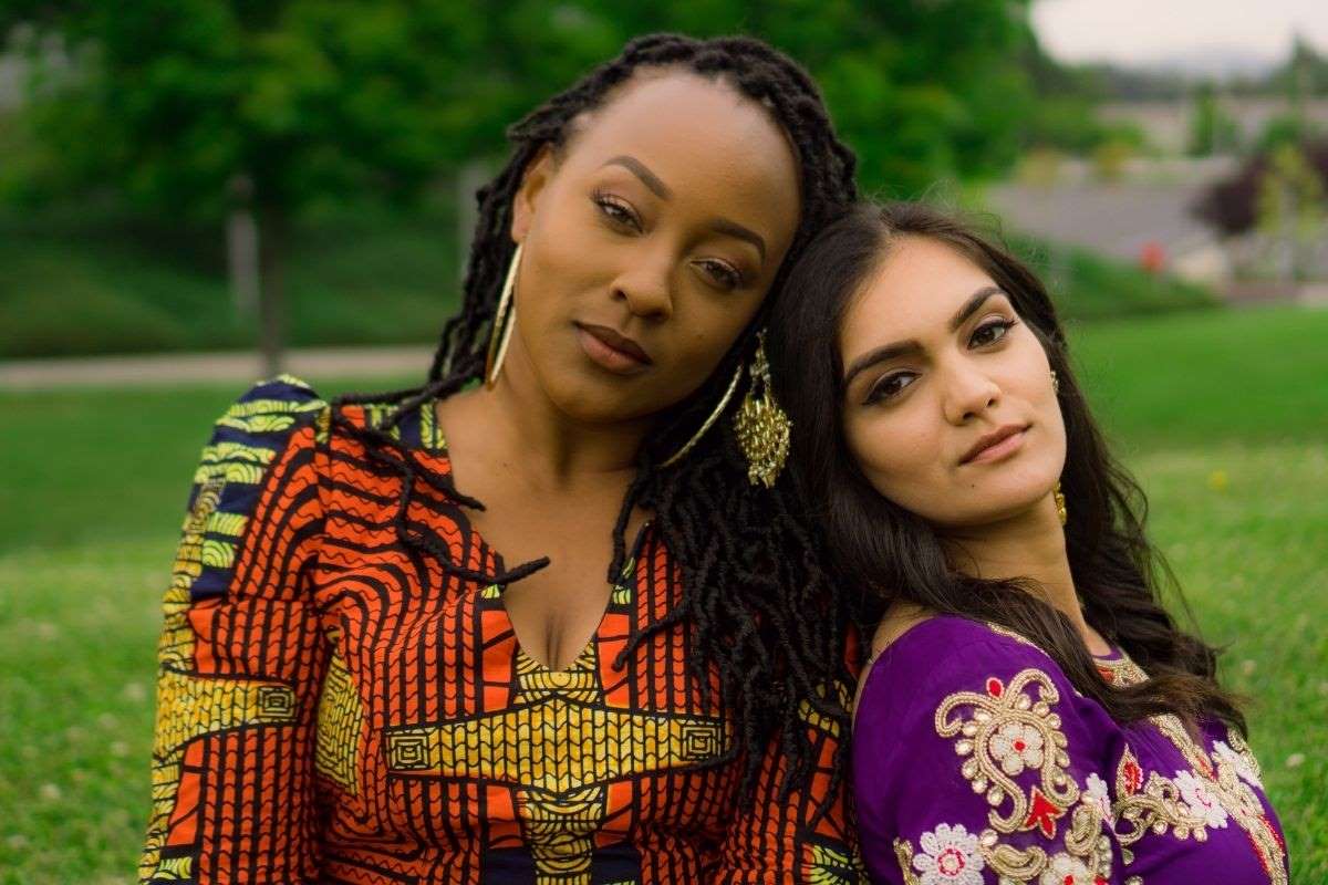 two women in colorful dresses and gold jewelry stand back to back looking at the camera