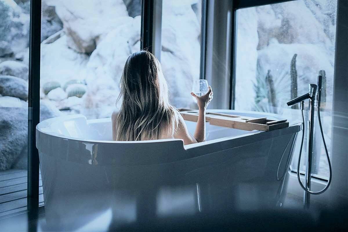 a woman sits in a bathtub with a glass of wine looking out windows at large bounlers