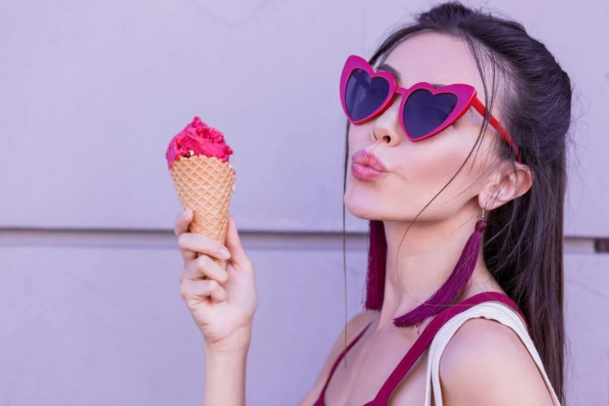 young woman in big heart shaped sunglasses and long dangling earrings looks flirtyand eats an ice cream cone