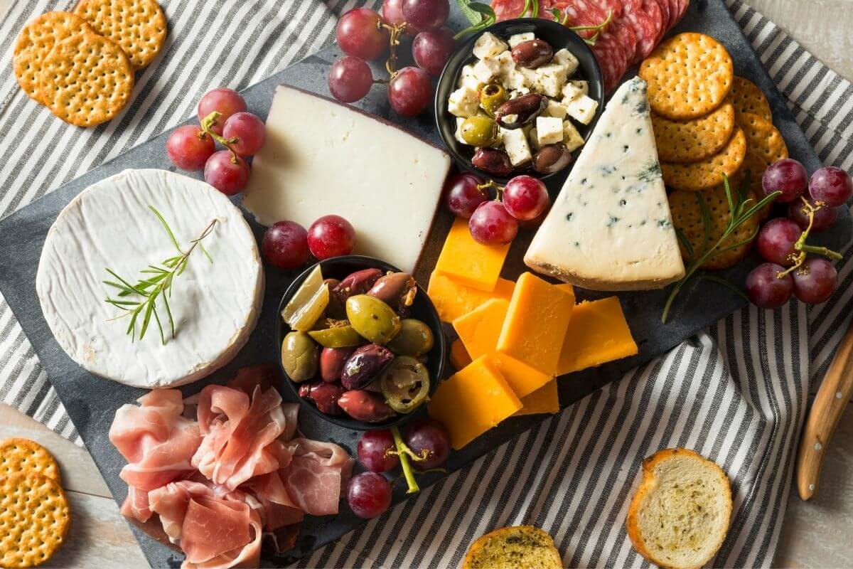 black charcuterie board with cheeses crackers fruits veggies and olives on a black and white striped placemat