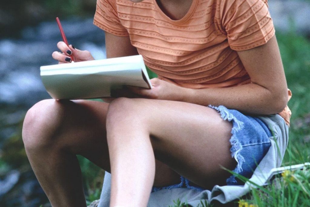 a woman sits outside on the grass writing a letter to her younger self