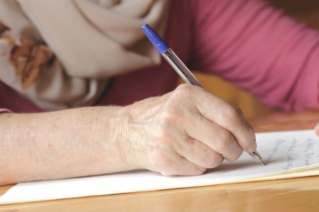 a woman with a scarf on sits at a wooden desk writing a letter