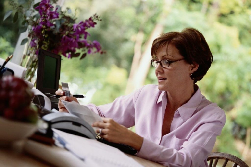 a woman sits at a table and writes a letter to her younger self with a vase of purple flowers next to her