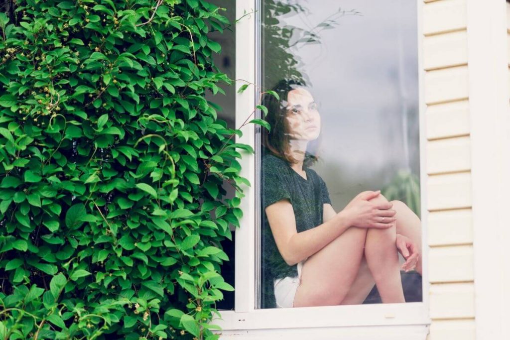 a young women sits on a window ledge looking outside with a look for worry on her face as she contemplates the struggles of life