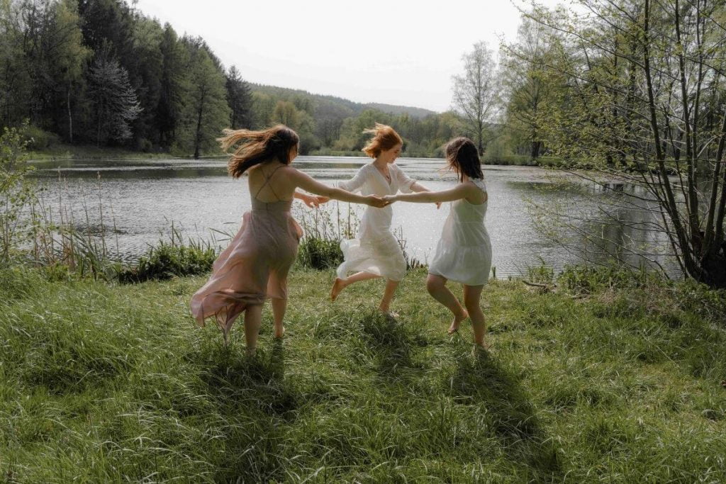 three young women dance in a circle holding hands along the edge of a lake with green hills in the background