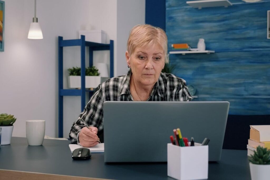 middle aged woman sits at a laptop with a pen in her hand staring blankly at the screen like shes overwhelmed