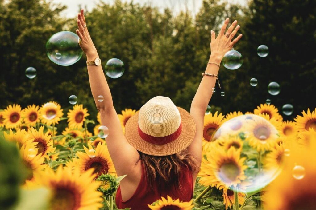 a woman in a red dress and a had with a red ribbon throws her hands up in the air in happiness in a sunflower field to show lets choose joy