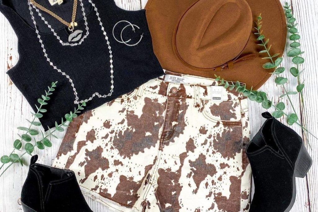 cow designed jean shorts black boots brown cowgirl hat black tank top silver earrings and necklaces laying on a white washed wooden background
