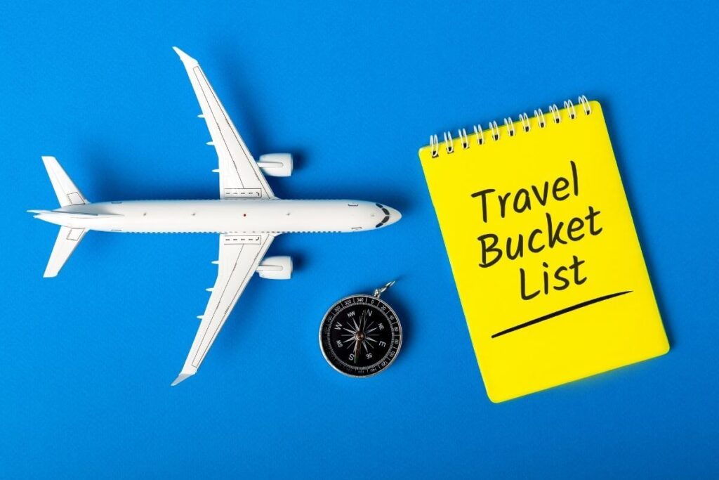 a white toy airplaince a black compass and a yellow travel bucket list notebook on a blue flat lay display