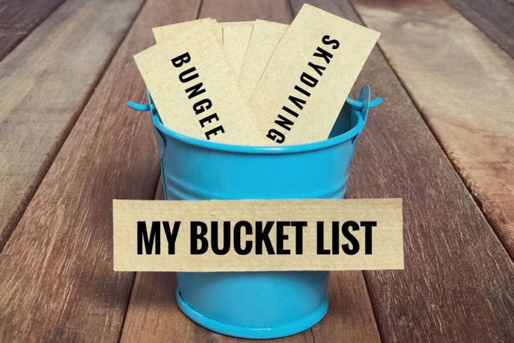a blue metal bucket sits on wood planks and has bucket list ideas printed on cards in it with my bucket list on the outside