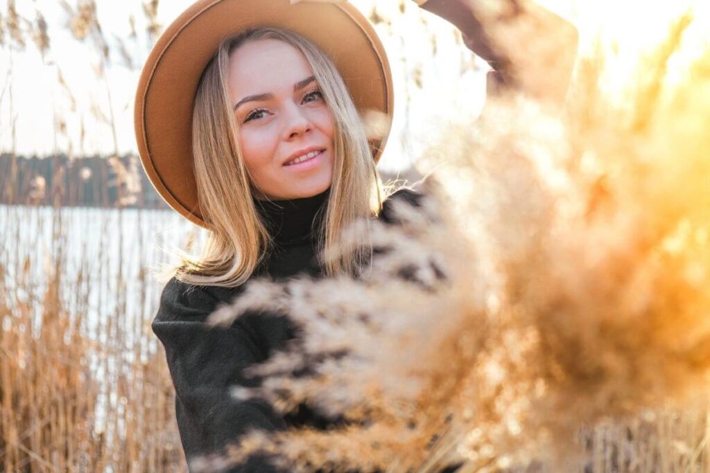 a young blonde woman wearing a tan hat stands in tall water grass along the shoreline and savors a slow living style