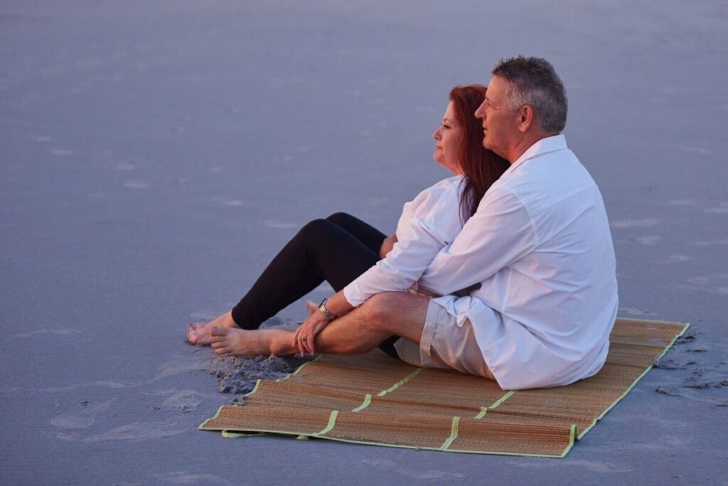 a middle aged man and woman sit comfortably and cozy on a mat on the beach demonstrating a slow living lifestyle
