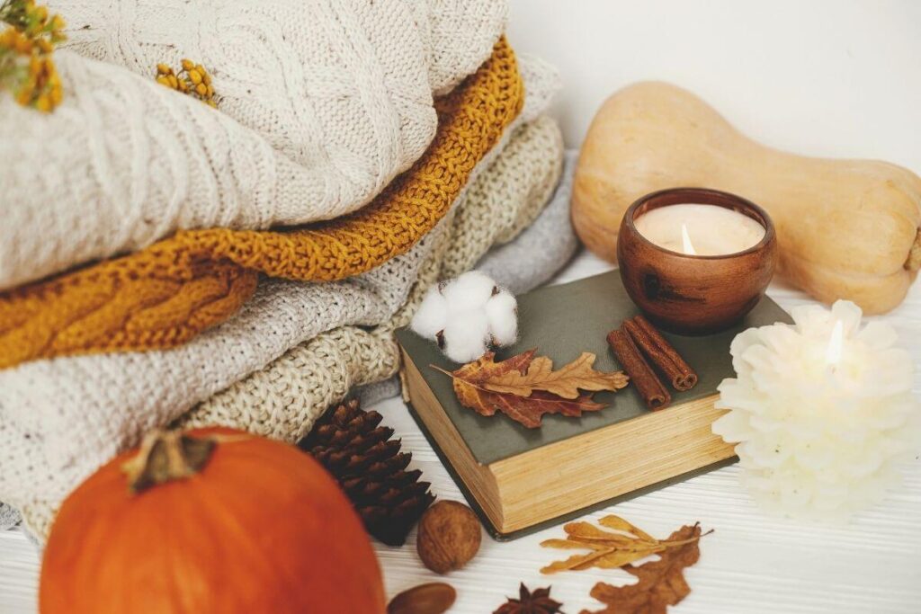 warm cozy sweaters and blankets folded next to a pumpkin pinecones walnuts leaves an old book a candle and a gourd in warm shades of beiges and crisp fall colors