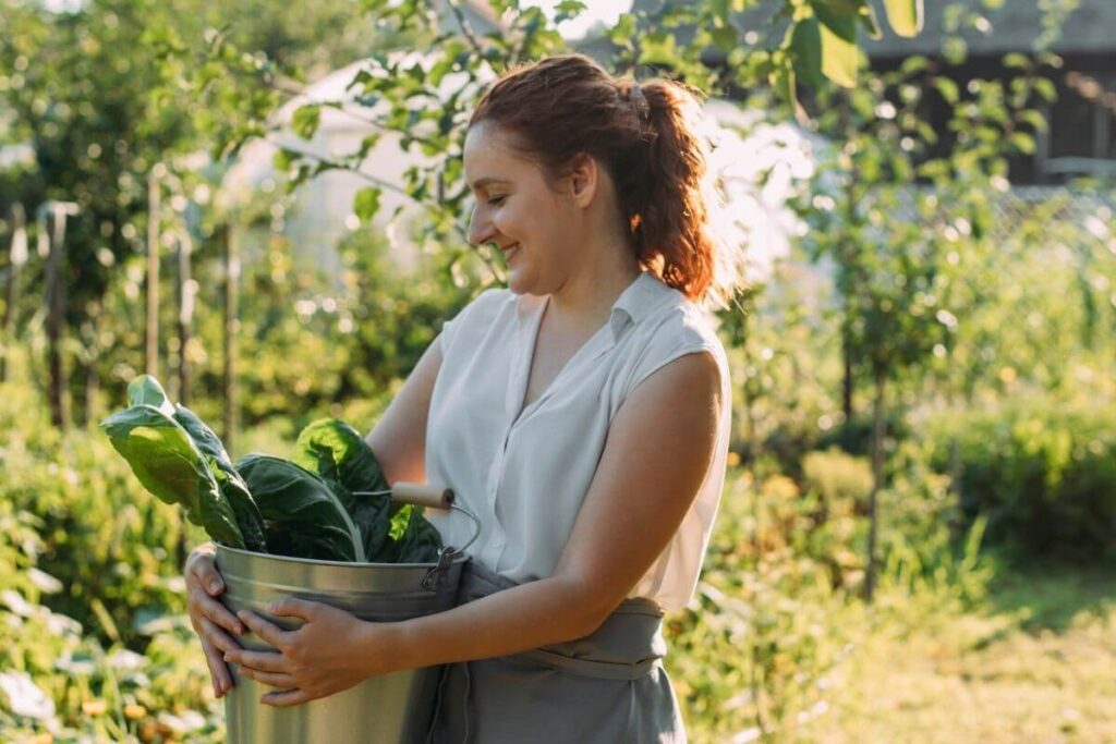 a woman carries a bucket of green leafy vegetables she has picked from a nearby garden and smiles happily about her slow living lifestyle