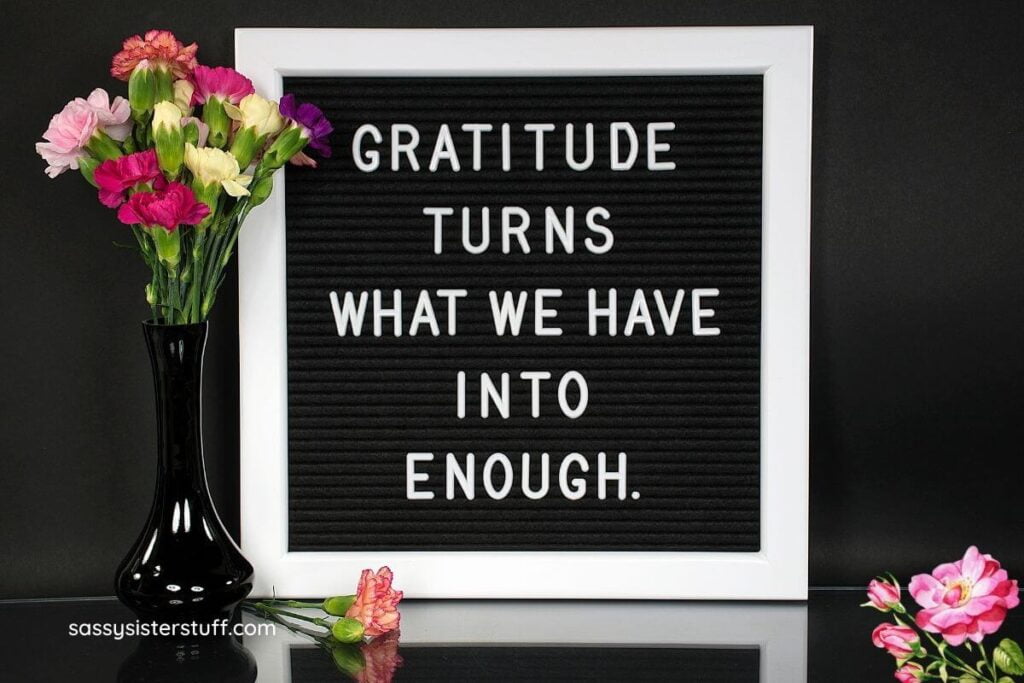 black background with a black and white frame that says gratitude turns what we have into enough plus a black vase of flowers and flowers laying on a black table