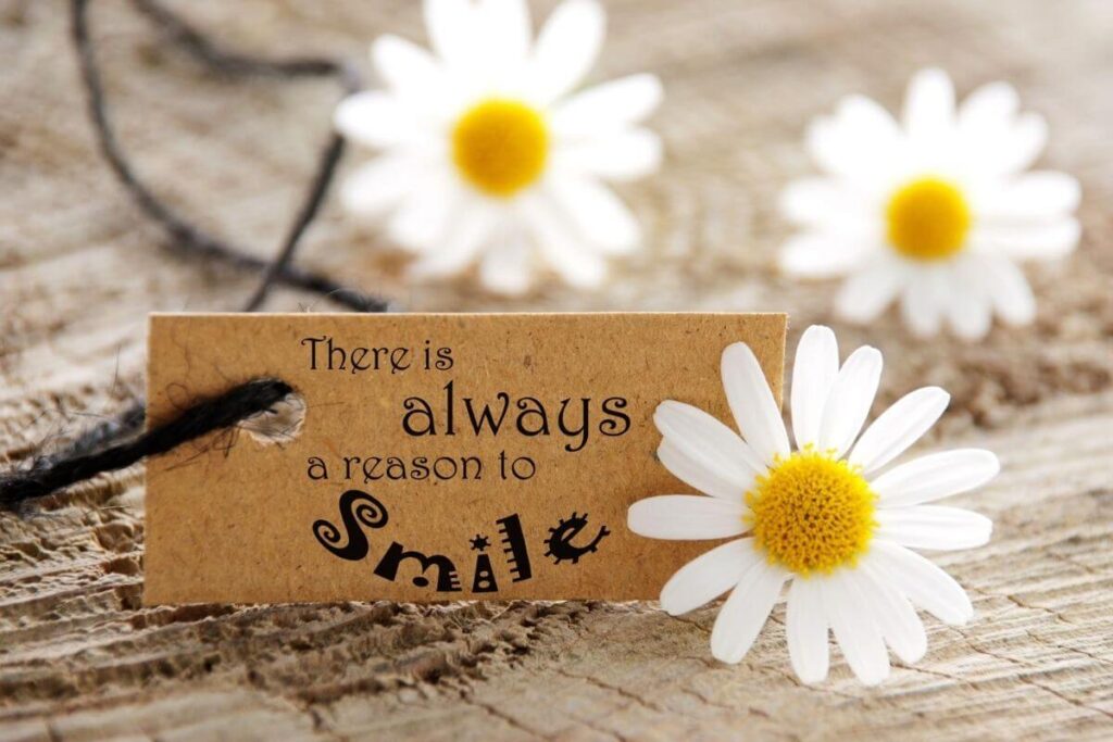 white daisies laying on sandy ground and a tag that says there is always a reason to smile