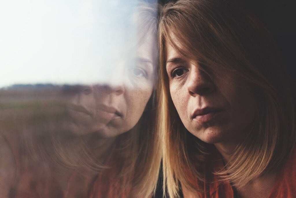a depressed female with shoulder length hair leans against a window staring into space while he reflection glares back as if defeated by an adult bully