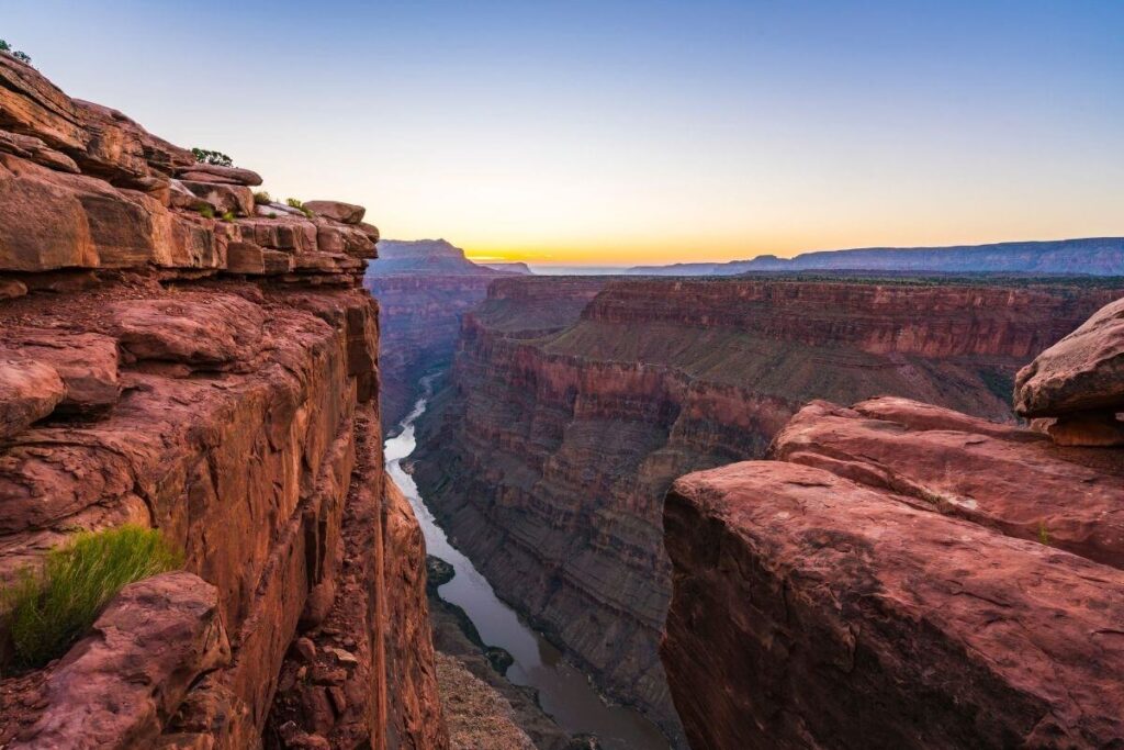 an image of the grand canyon walls with the river flowing at the bottom and a beautiful blue and yellow sky above shows one of the most adventurous bucket list locations in america
