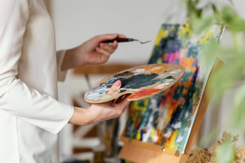 a woman holds a paint tray of paints in front of a canvas that is she painted in abstract designs and various colors