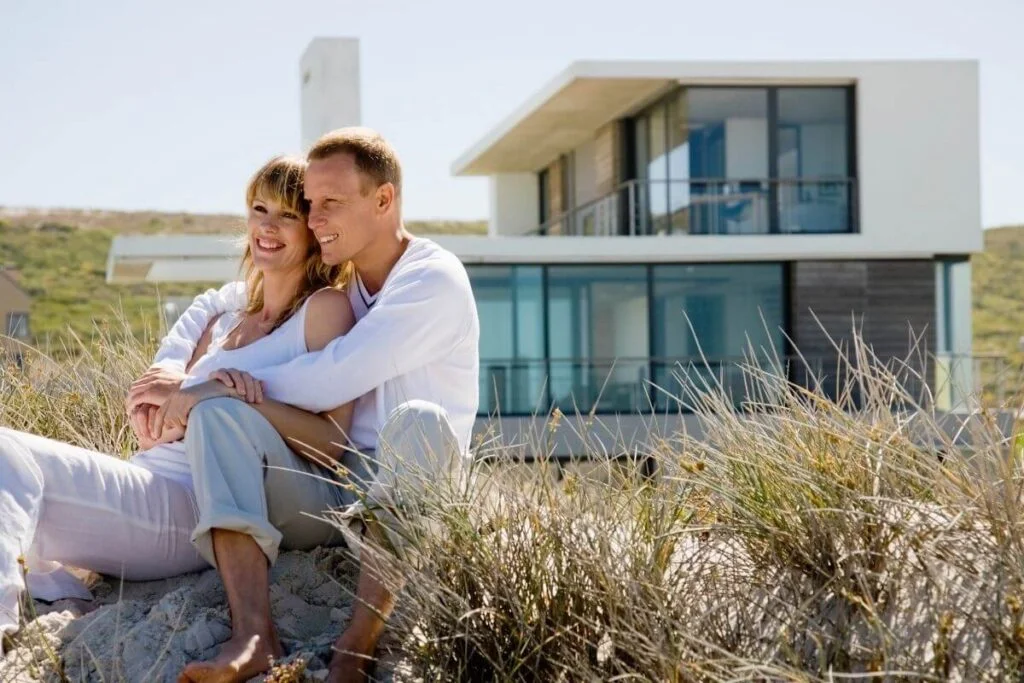 young man and woman sit together in water grasses at the beach in front of a beautiful new beach house sitting on a hill behind them