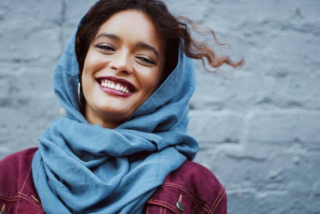 beautiful ethnic woman with a blue wrap around her head and bright pretty make-up stands against a pale blue brick wall smiling at the camera