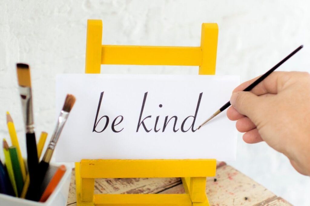 a hand from the right side of the image paints the words be kind on a small piece of cardboard sitting on a small yellow easel with colored pencils and paint brushes next to it