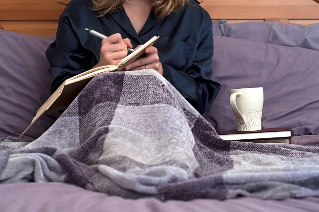 top view of woman sitting in bed writing in her journal with a mug and some books sitting next to her
