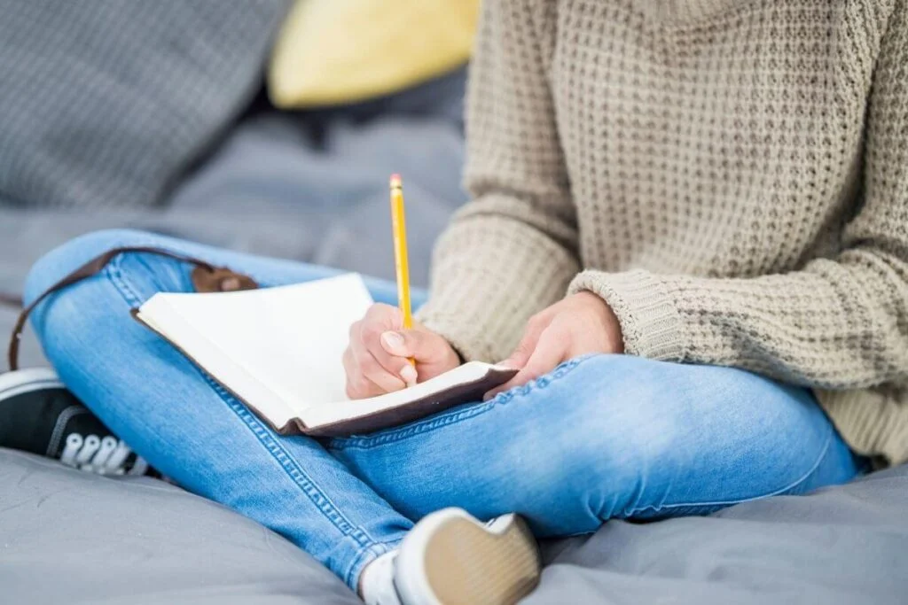 partial view of woman sitting on a comfy blanket using journaling prompts for healing as she writes in her journal