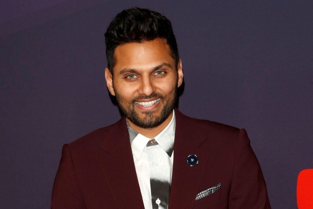 a close up image of Jay Shetty dressed in a burgundy suit standing against a purplish black background