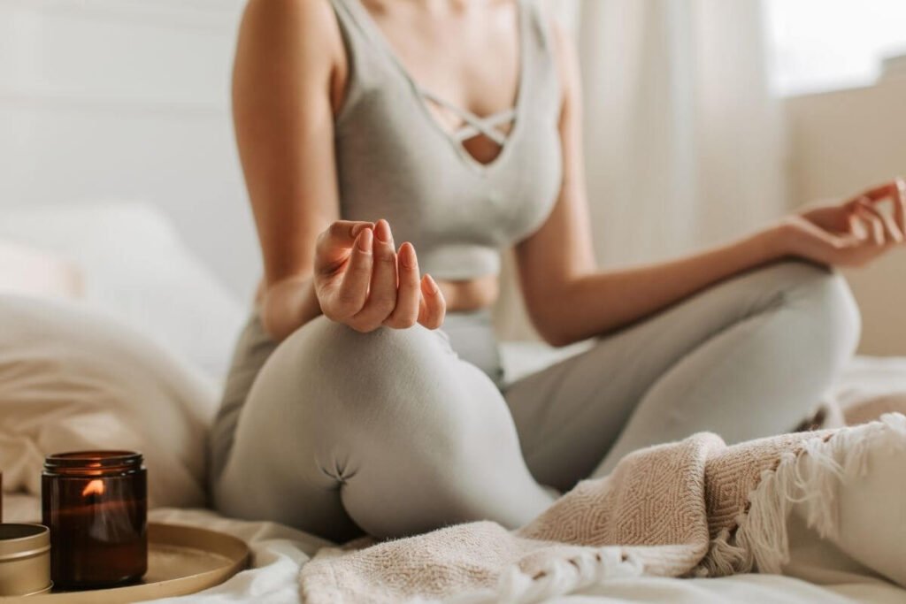a close up of a female going meditation with her legs crossed and her hands resting on her knees and a candle sitting next to her plus other meditation room essentials like a blanket and pillow