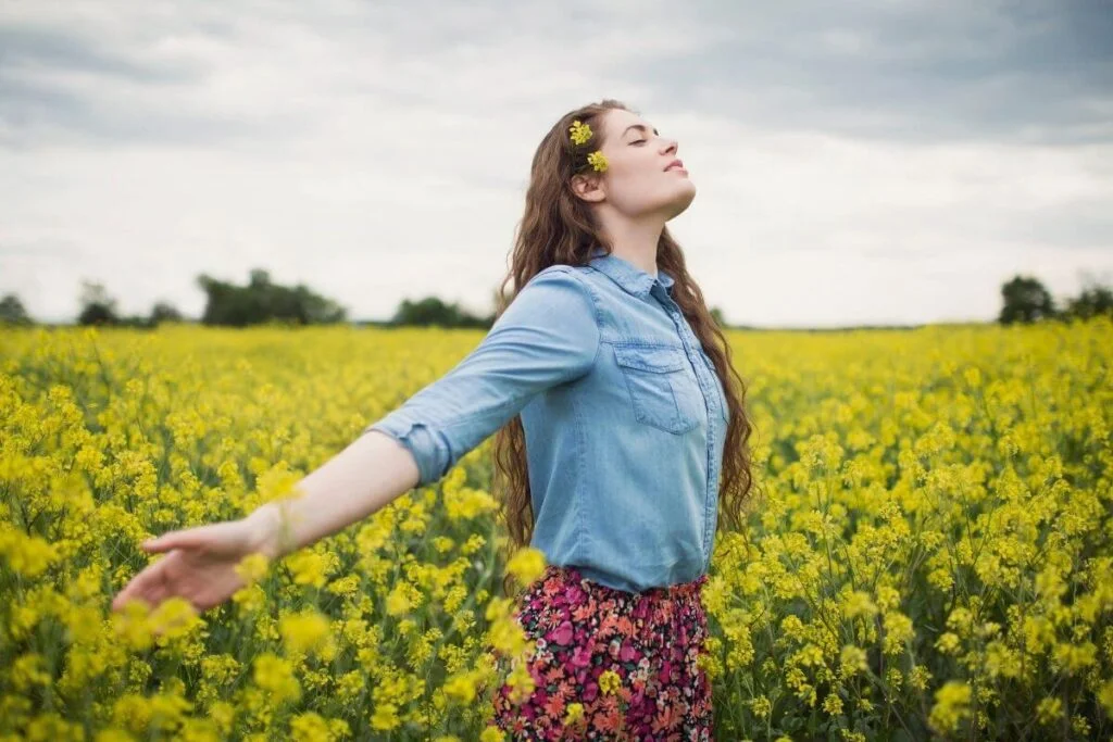 a young happy woman with long dark brown hair stands in a yellow field of flowers on a beautiful sunny day as she mindfully enjoys the smells sights and sounds of the field