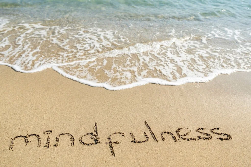 a calming close-up image of a beach shoreline with mindfulness written in the sand