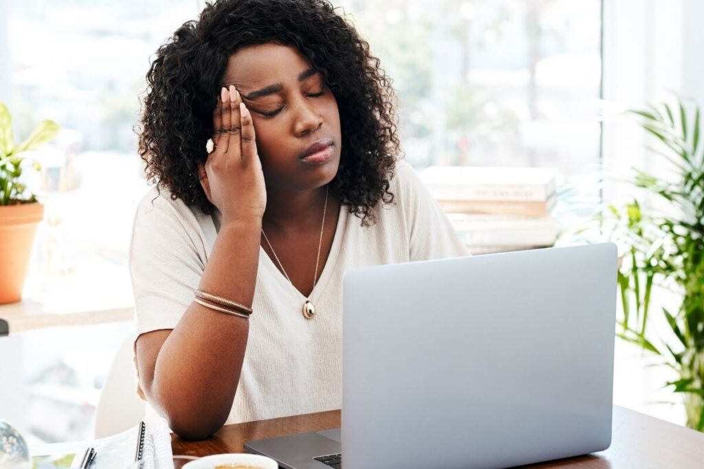 a woman sits at her desk in a sunny office with her hand on her head feeling frustrated using her laptop to search for personal growth tips for carerr burnout