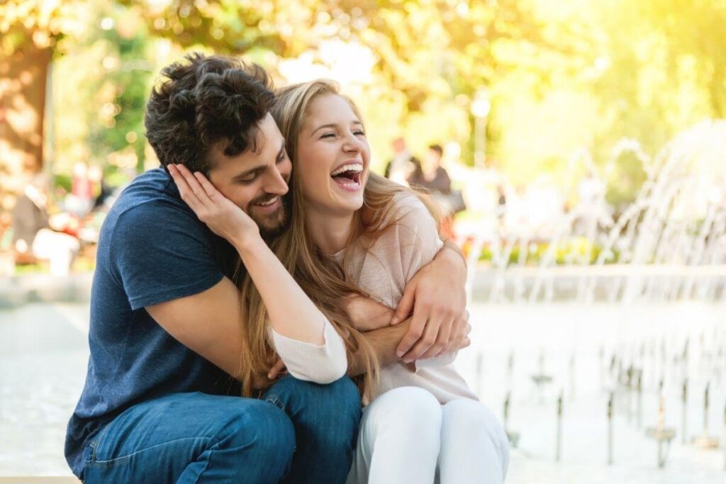 a young happy couple laughs and hugs as they sit outside on the edge of a water fountain on a sunny day as they work on creating a happy marriage