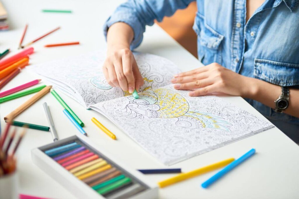 a women wearing a demin shirt is seen from a top view as she colors in an adult coloring book on a desk with a large selection of colored pencils