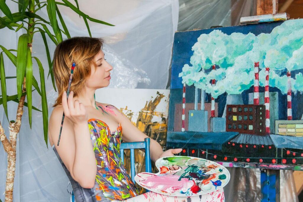 a young whimsical woman cover in bright paints on her top sits in a chair staring at a painting she is working on with a palette of paints in one hand and a paint brush in the other