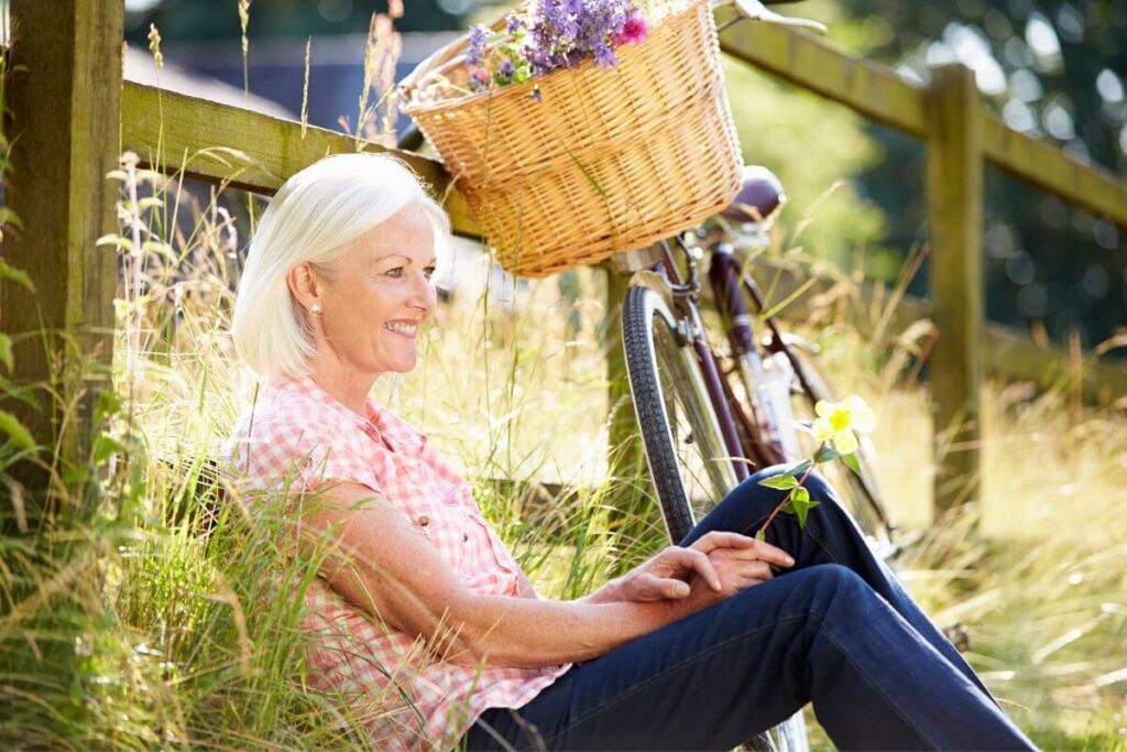 a middle aged blonde woman takes a break from her bike riding in the country and sits against a wooden fence smiling because she believes it's important to fall in love with taking care of yourself