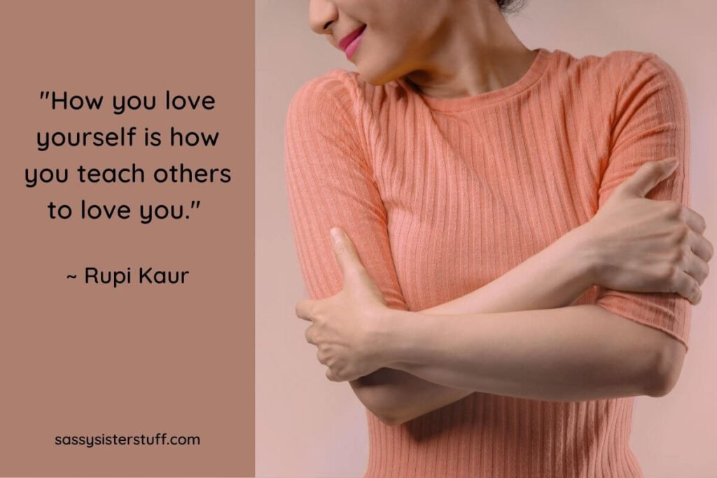 a young happy woman in a peach sweater hugs herself to show that self love isn't selfish and a self love quote from Rupi inspires her