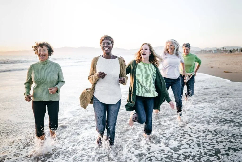 a group of middle aged women run in the waves along the shoreline for one of many unusual self care ideas they practice