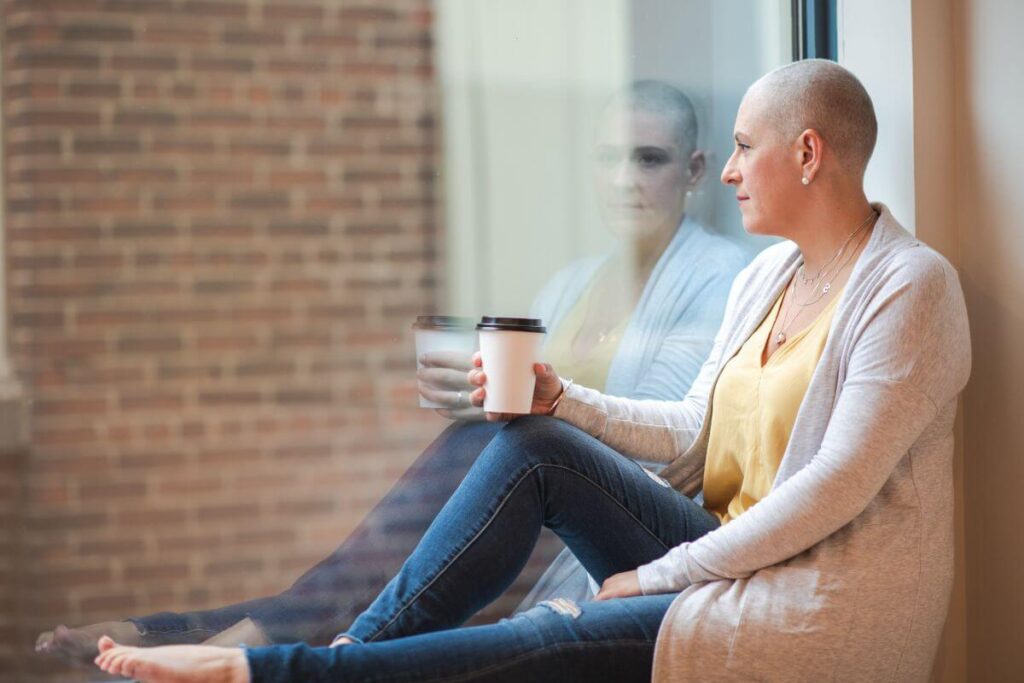 a pensive woman with a bald head sits on the ground looking outside a window things about how courageous she has been in her health challenge