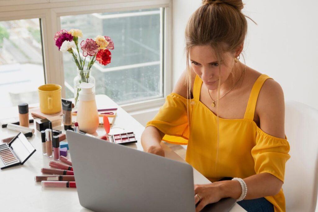 a blogger works on her laptop on a desk covered with assorted make up that she is writing about for women over 50