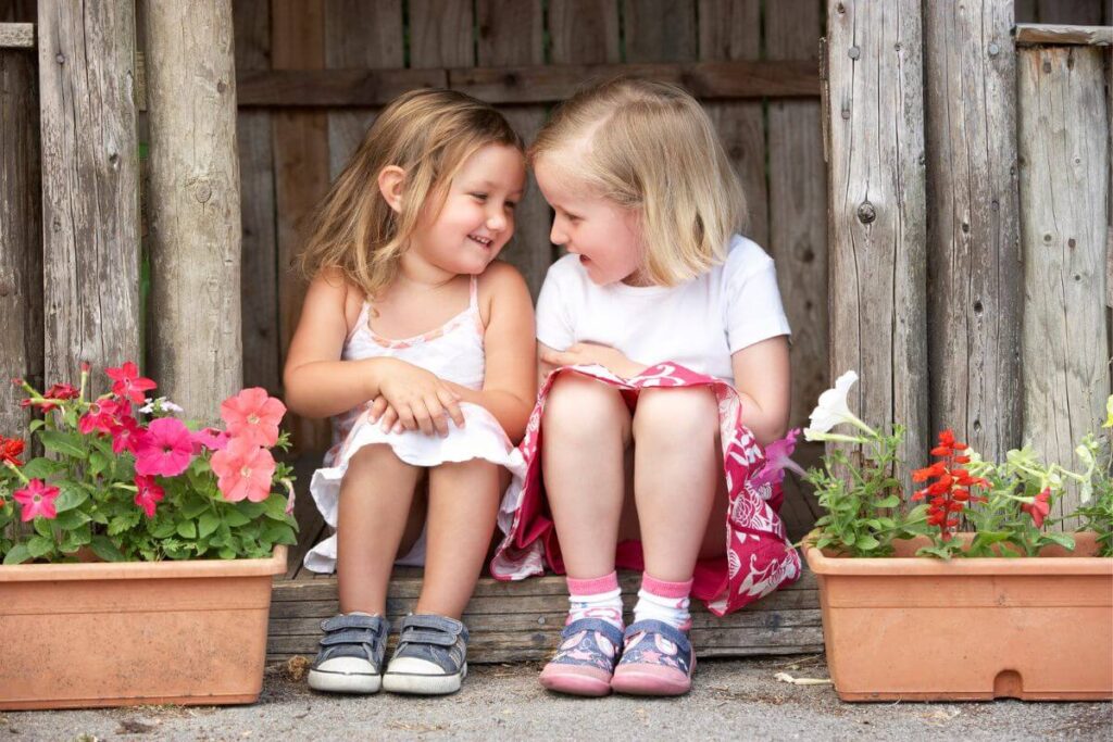 two little girls sit on a wooden porch next to each other laughing together