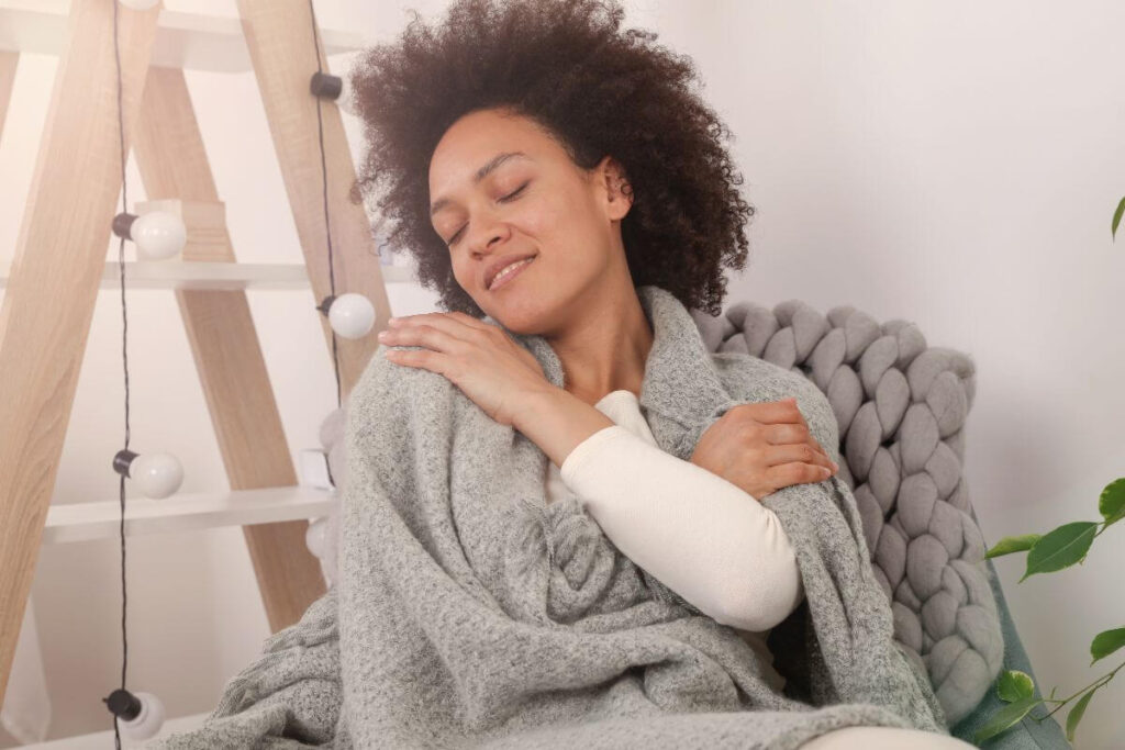a young woman with brown hair sits in a plush chair wrapped in a blanket and embraces herself in a sign of self love