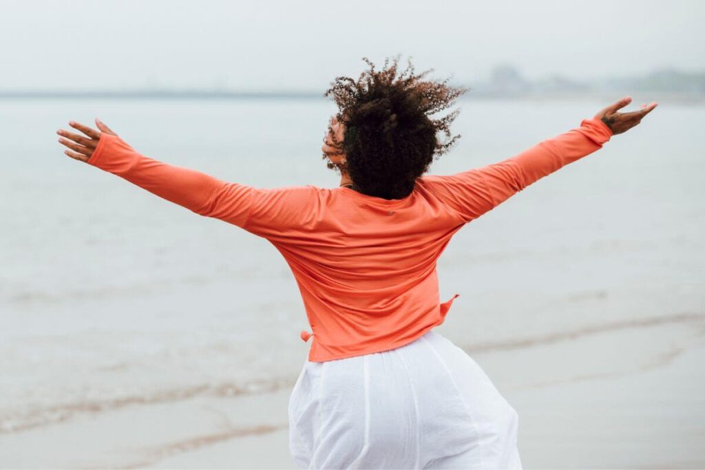 a very happy woman throws her arms in the air in happiness as she walks along a beach