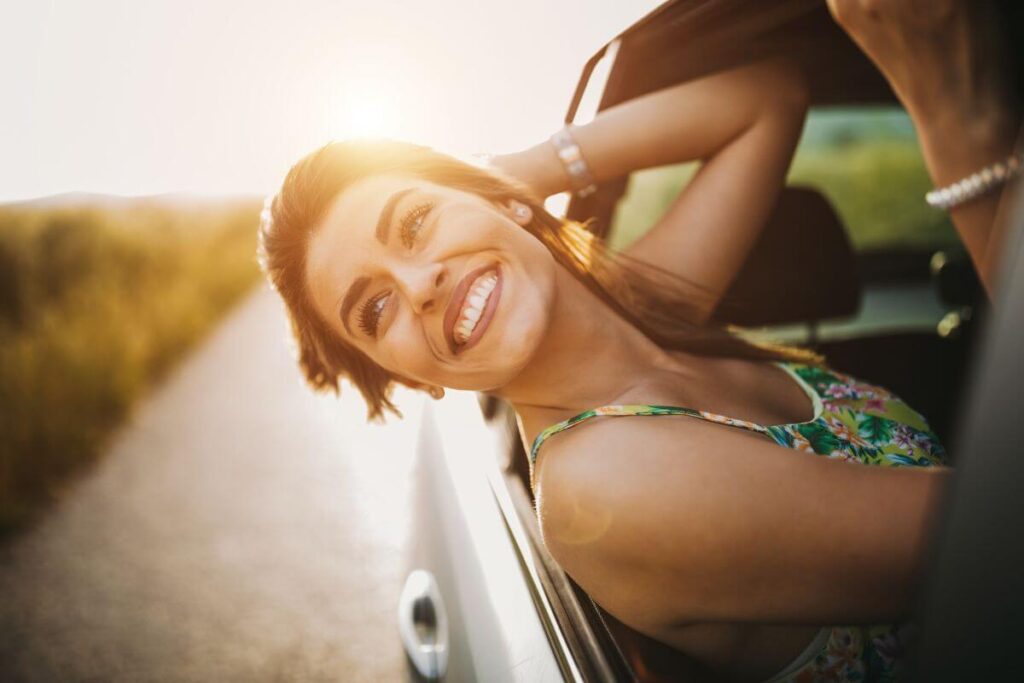 a young woman rides in a car and hangs her head out of the window in fun as she lives her best life