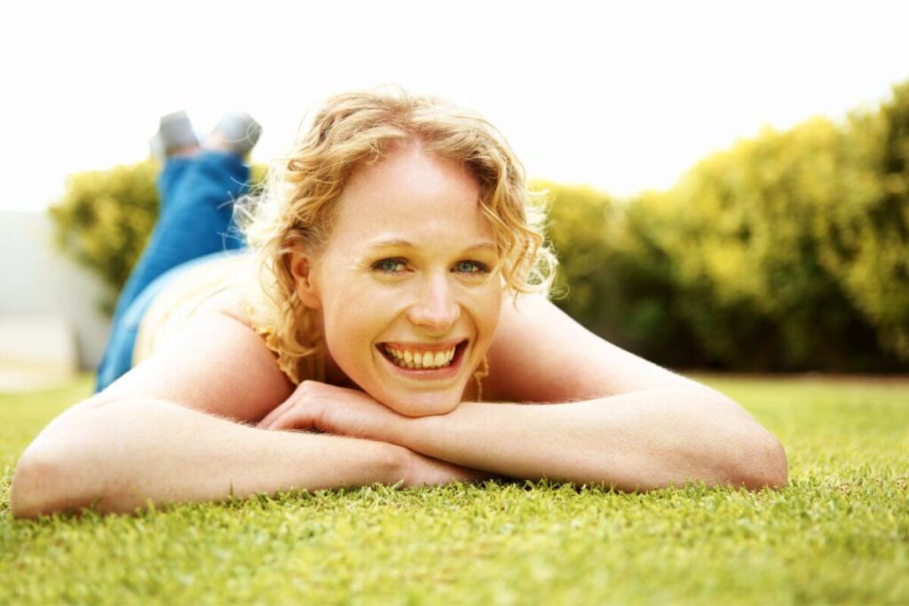 a very happy woman lays on her stomach in the grass and smiles at a camera because she is living her best life on a sunny day