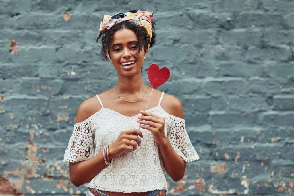 beautiful young woman stands against a brick wall holding a red heart to show she prioritizes herself