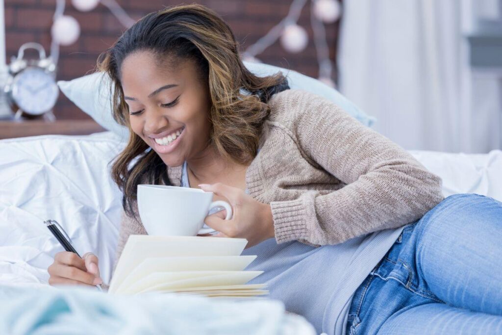 a beautiful young dark skinned woman in jeans and a tan sweater lays across her bed with a cup of coffee using wellness journal ideas and prompts for her daily journaling